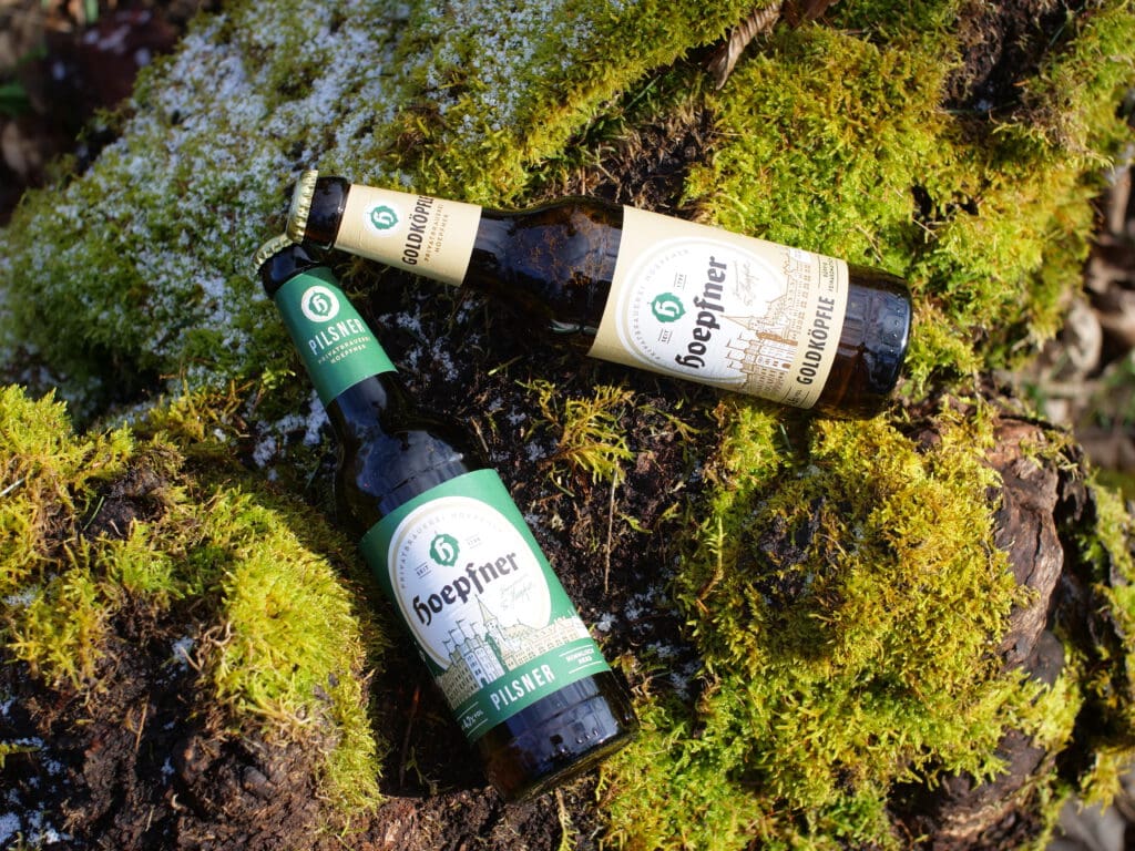 Zwei Hoepfner Privatbrauerei Flaschen auf moosbedecktem Baumstamm.